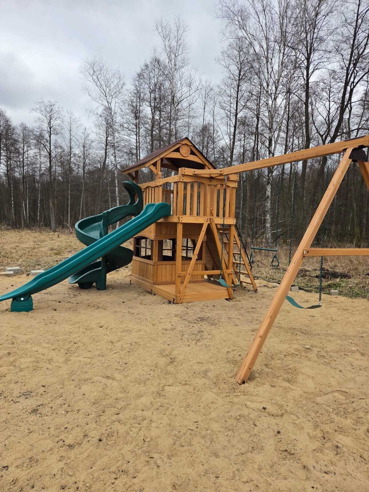 Estructura de madera para juegos con tobogán y columpios en una zona arenosa, con árboles al fondo.