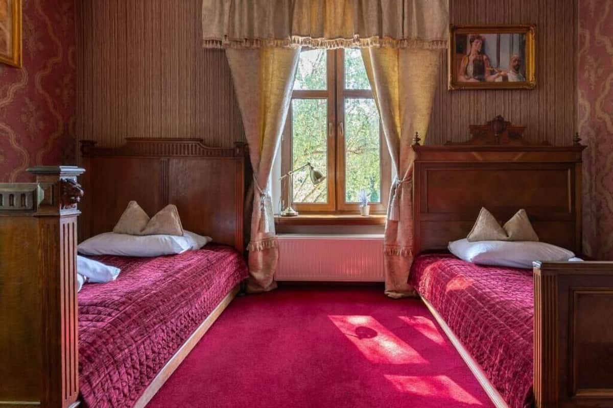 Una habitación con dos camas de madera, una ventana con cortinas, un radiador y un cuadro en la pared.