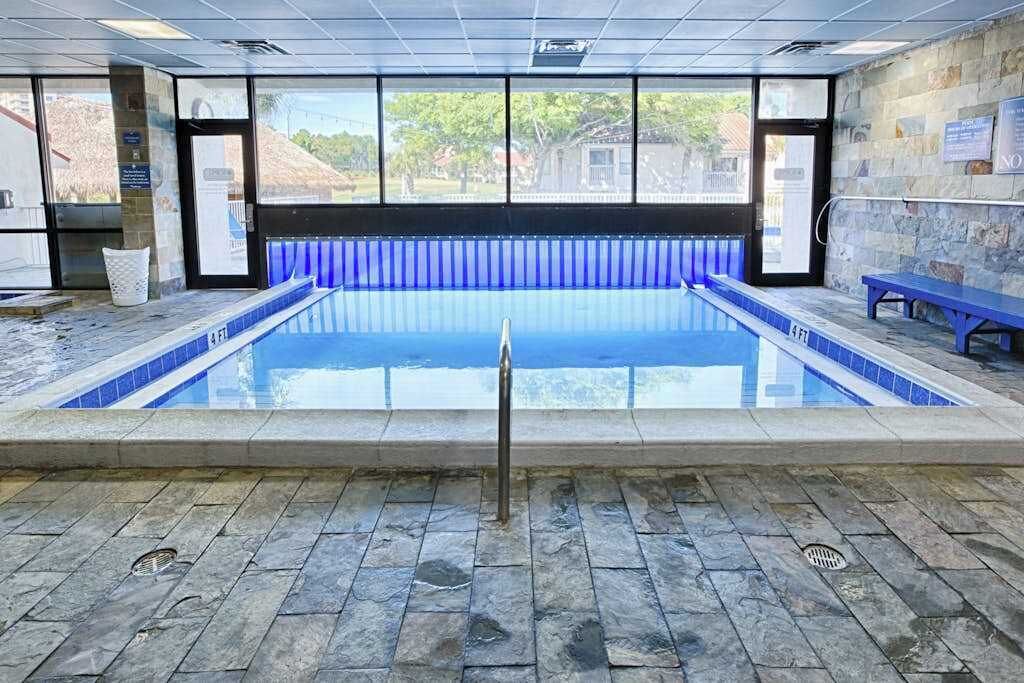 Piscina interna com água azul cristalina, piso de pedra e vista para o exterior através de grandes janelas.