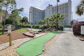 A miniature golf course with a green putting surface, a numbered sign, and a backdrop of tall buildings and palm trees. - Spacious Coastal Escape for Family Getaways (Miramar Beach)