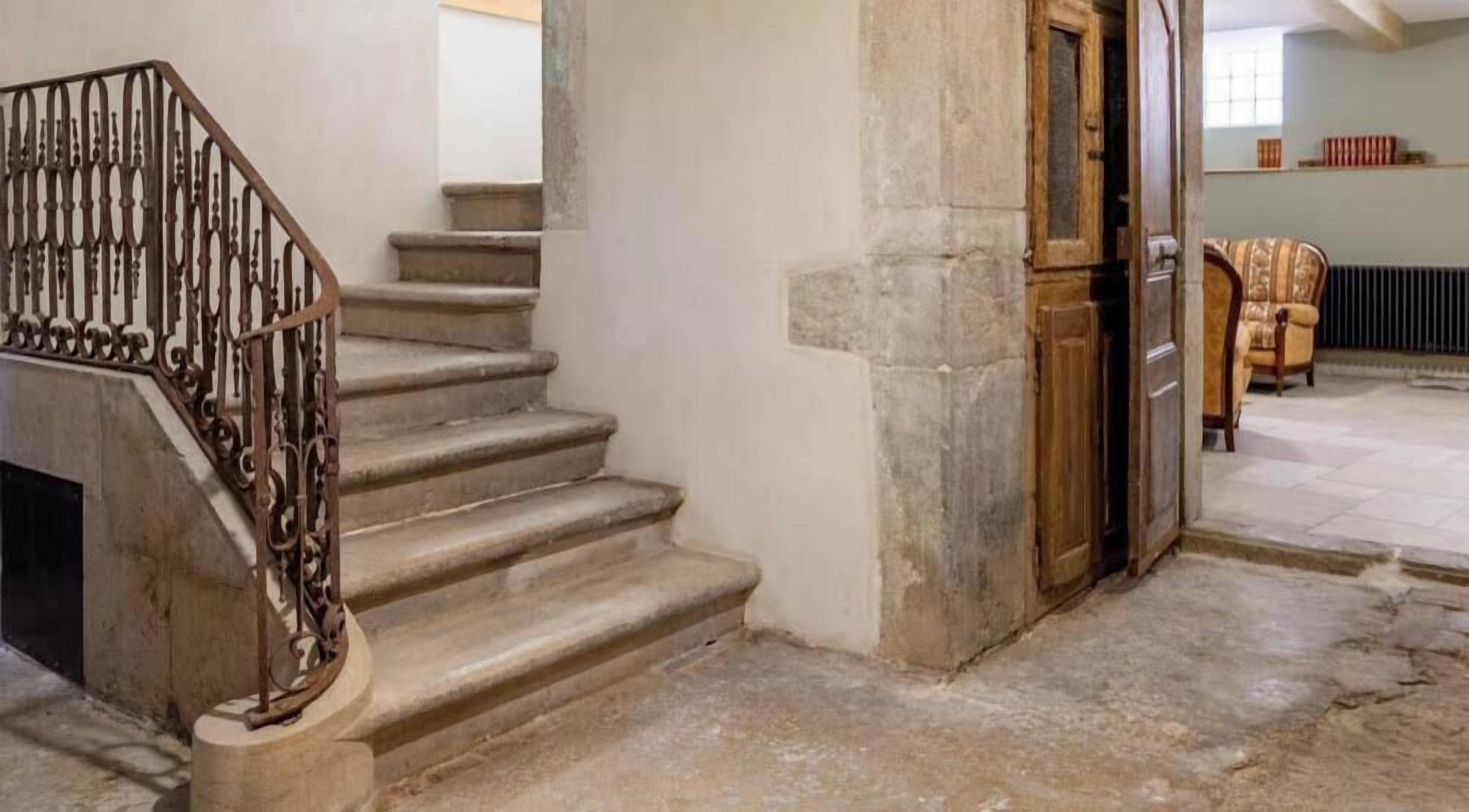 Eine Treppe mit schmiedeeisernem Geländer führt zu einem Raum mit einer Steinwand und einer Holztür.