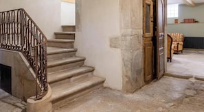 A stone staircase with a wrought iron railing, a wooden door, and a chair in a room with stone walls and a tiled floor. - Stendhal House - Le Belvédère (LES ECHELLES)