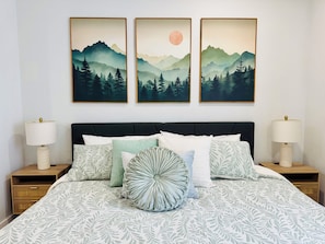 A bedroom with a bed, pillows, a nightstand, a lamp, and three framed landscape paintings on the wall. - Gorgeous two-bed suite + bonus media room and sleeping loft in updated duplex (Idaho Falls)