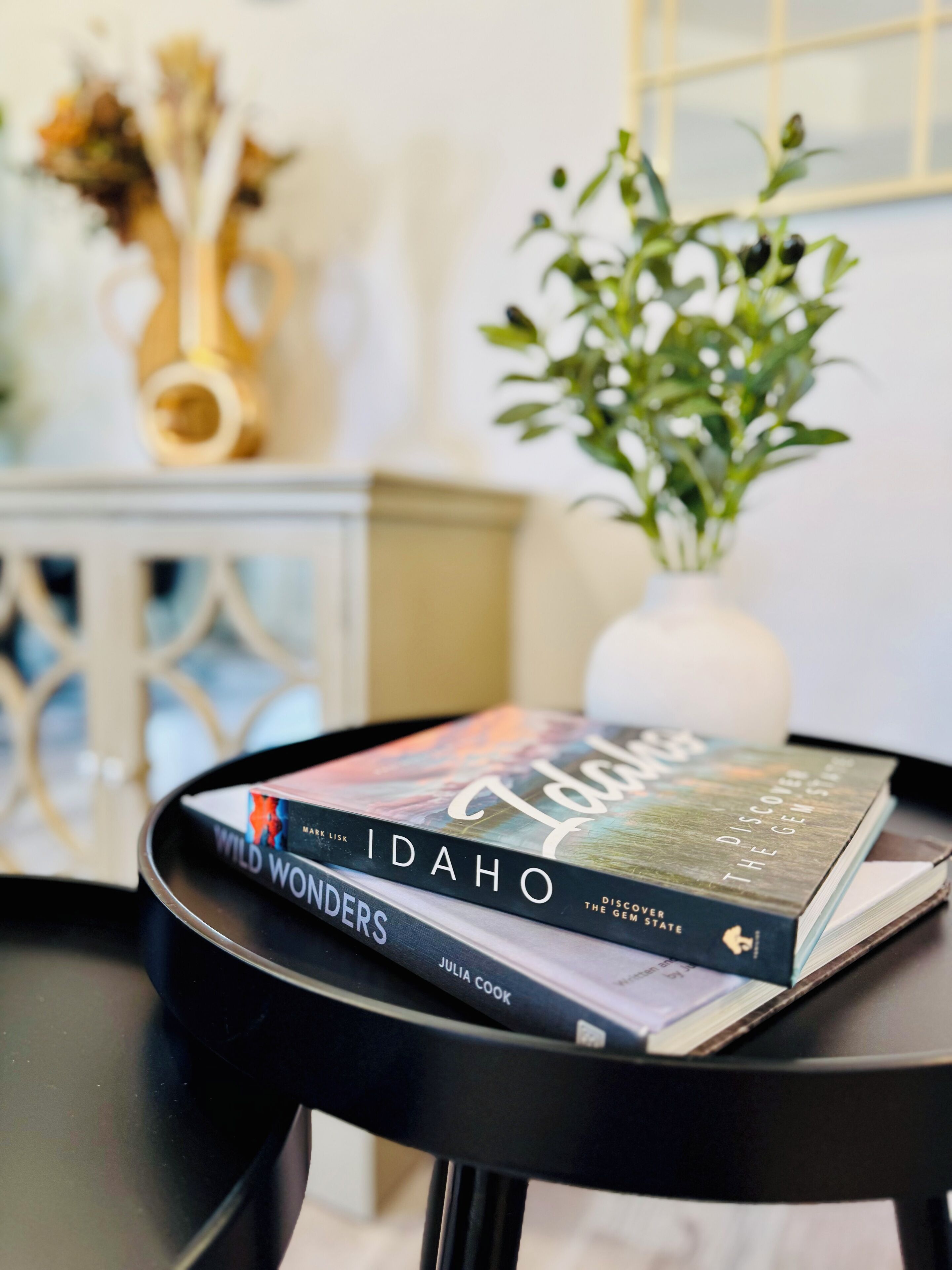 Ein Stapel Bücher auf einem schwarzen Tisch, im Hintergrund eine weiße Vase und eine Pflanze.