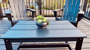 Una terraza de madera con una mesa y dos sillas Adirondack, una planta en maceta sobre la mesa, y vistas a una calle residencial y árboles.