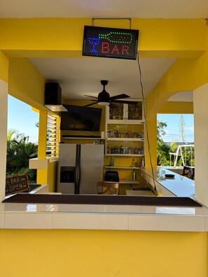 Eine Bar mit einer Neonreklame, einem Deckenventilator und Blick ins Freie.