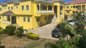 A yellow multi-story building with a central staircase, balconies, and a covered driveway. - Welcome to Spring Dash,2BR POOL&BEACH ACCESS+VIEW,FREE PARKING,BAR&Grill, &AC (St Ann)