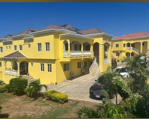 A yellow multi-story building with a central staircase, balconies, and a covered driveway. - Welcome to Spring Dash,2BR POOL&BEACH ACCESS+VIEW,FREE PARKING,BAR&Grill, &AC (St Ann)