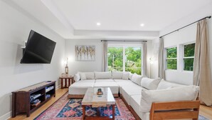 A modern living room with a sectional sofa, a flat-screen TV, a coffee table, and a large window with curtains. - Modern 3BR San Marco Villa (Jacksonville)