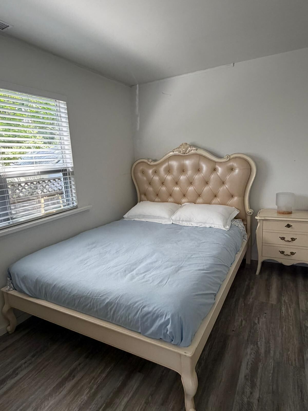 Un dormitorio con una cama, una mesita de noche, un ventanal con persianas y suelo de madera.