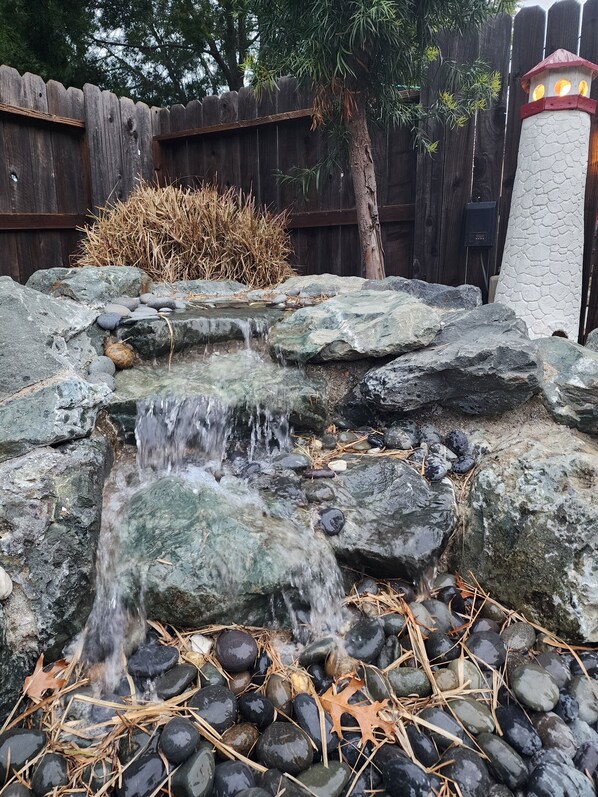 Ein kleiner Wasserfall mit Steinen und Kieselsteinen, umgeben von trockenem Gras und einem Holzzaun.