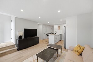 A modern living room with a sofa, a coffee table, a TV, and a kitchen area with cabinets and a refrigerator. - The Calgary Stopover (Calgary)