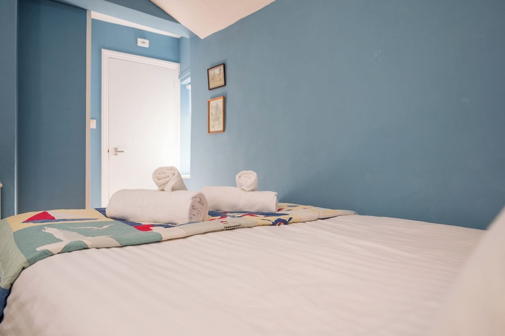 A bedroom with a bed, two rolled towels, a door, and a wall with two framed pictures.