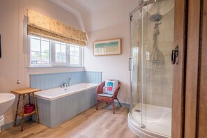 A bathroom with a bathtub, a shower, a window with a view, a wicker chair, and a wooden floor. - Four Gables, Lakeside Avenue, Thorpeness (Thorpeness)