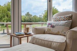 Ein Zimmer mit einem gepolsterten Sessel, einem kleinen Tisch mit einem Buch und einer Tasse Kaffee sowie Blick auf einen Garten durch große Fenster.