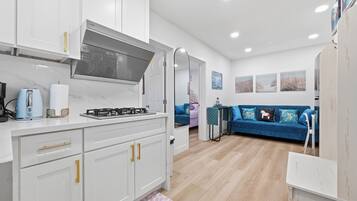 A modern kitchen with white cabinets, a gas stove, and a built-in oven. There is a blue sofa and a small table in the living area.
