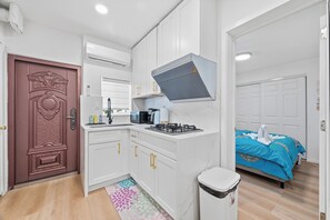 Une cuisine de taille réduite avec des meubles blancs, une cuisinière et un four à micro-ondes. Un tapis à motifs recouvre le sol et une porte au décor travaillé complète l’ensemble.