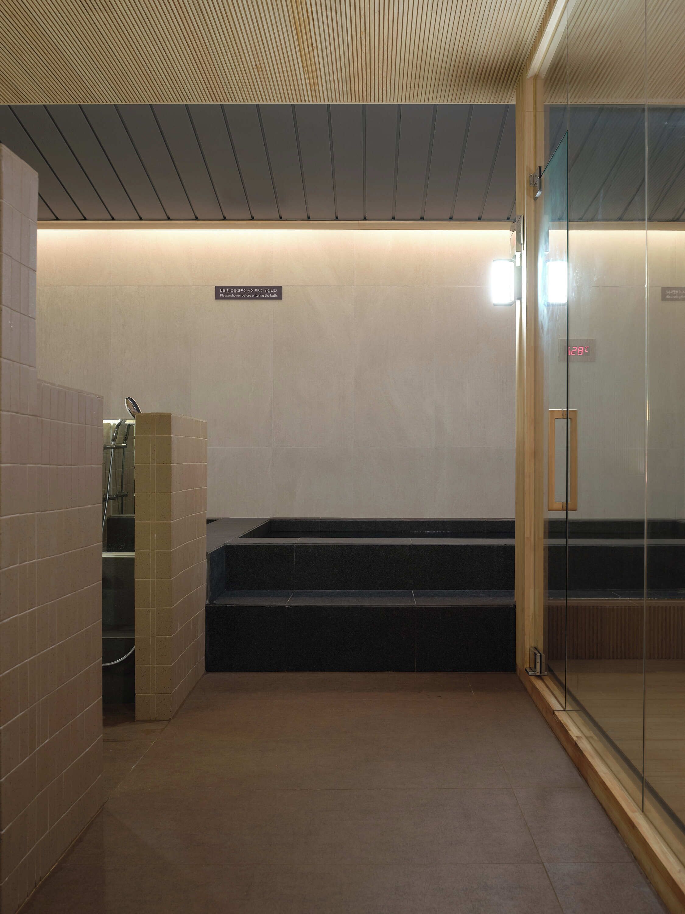 A modern bathroom with a glass partition, a sign indicating a shower before the bath, and a digital clock displaying 12:28.