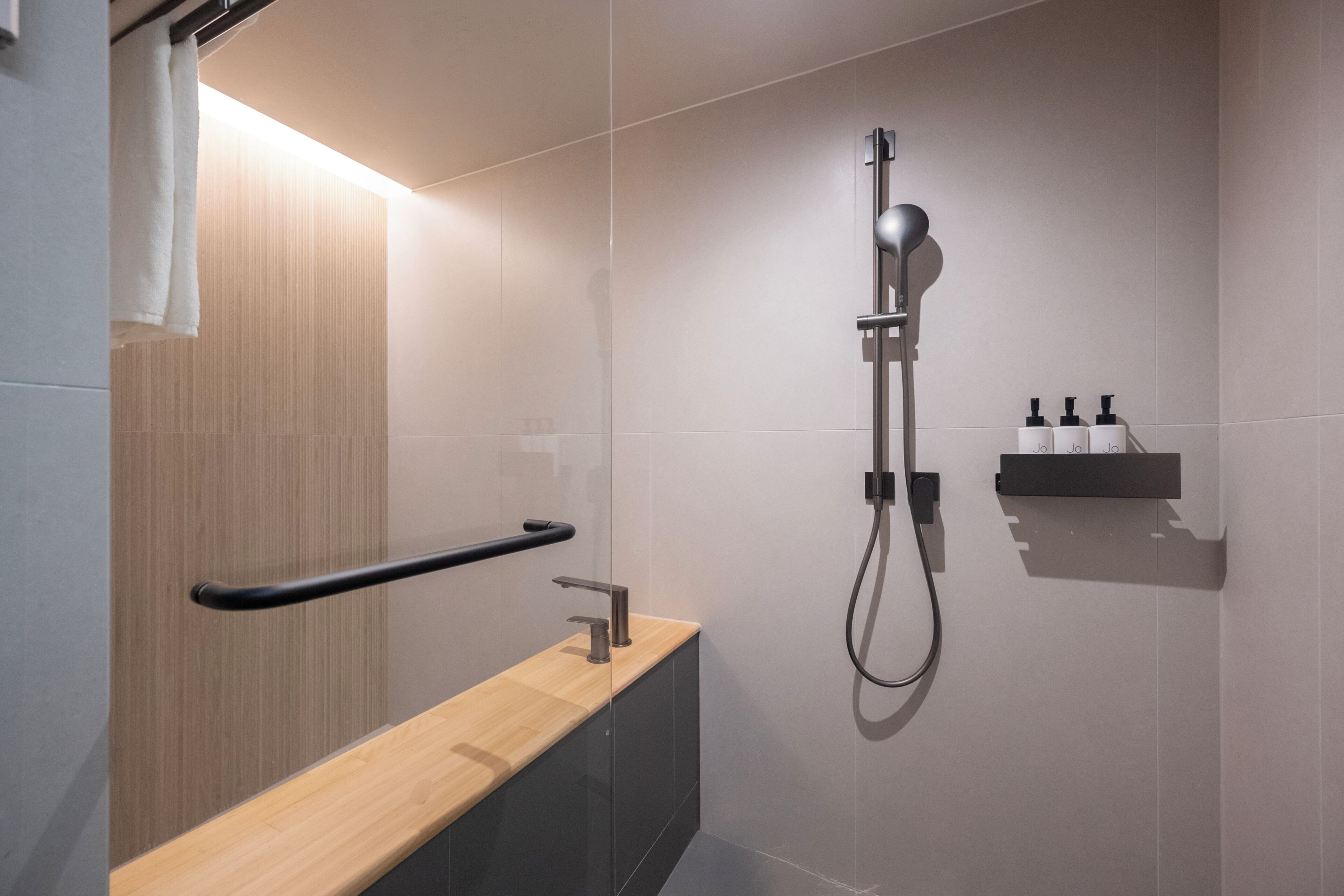 A modern bathroom with a wooden vanity, a towel rack, a shower with a handheld showerhead, and a soap dispenser on the wall.