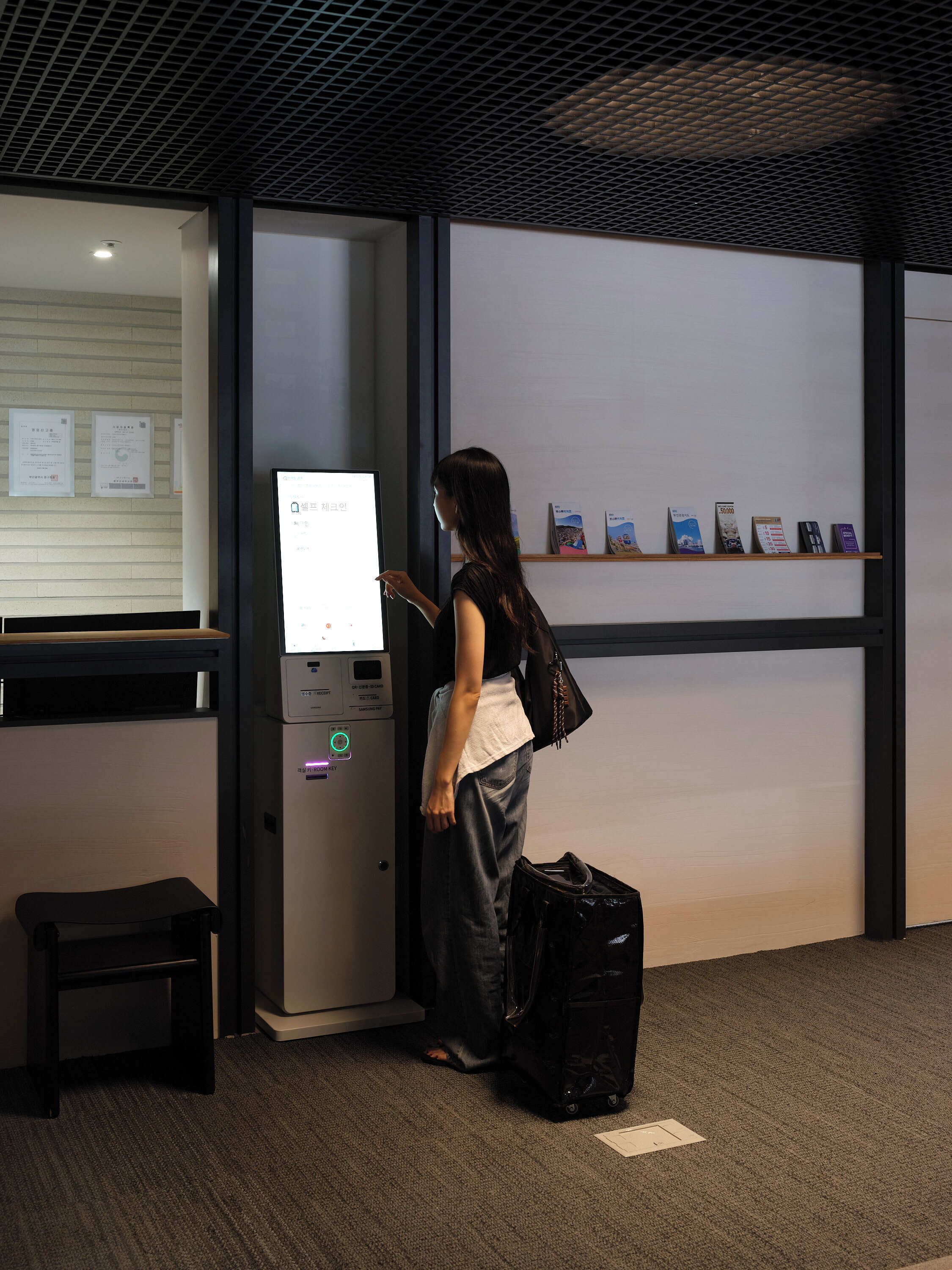 A woman standing next to a ticket kiosk with a digital display, a small desk with a chair, and a shelf with brochures.