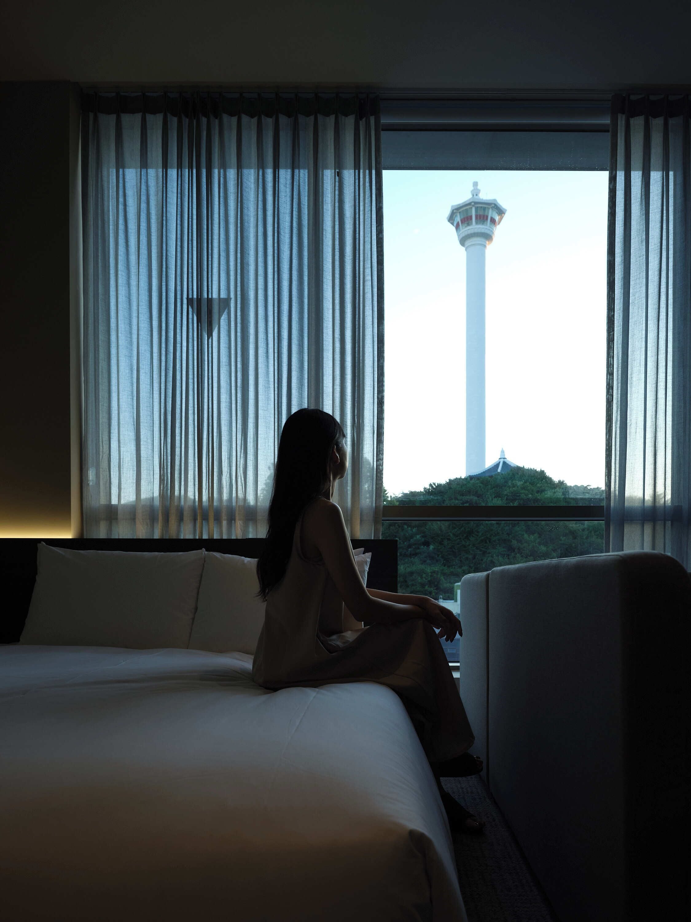 A hotel room with a bed, a chair, and a view of a tower through sheer curtains.