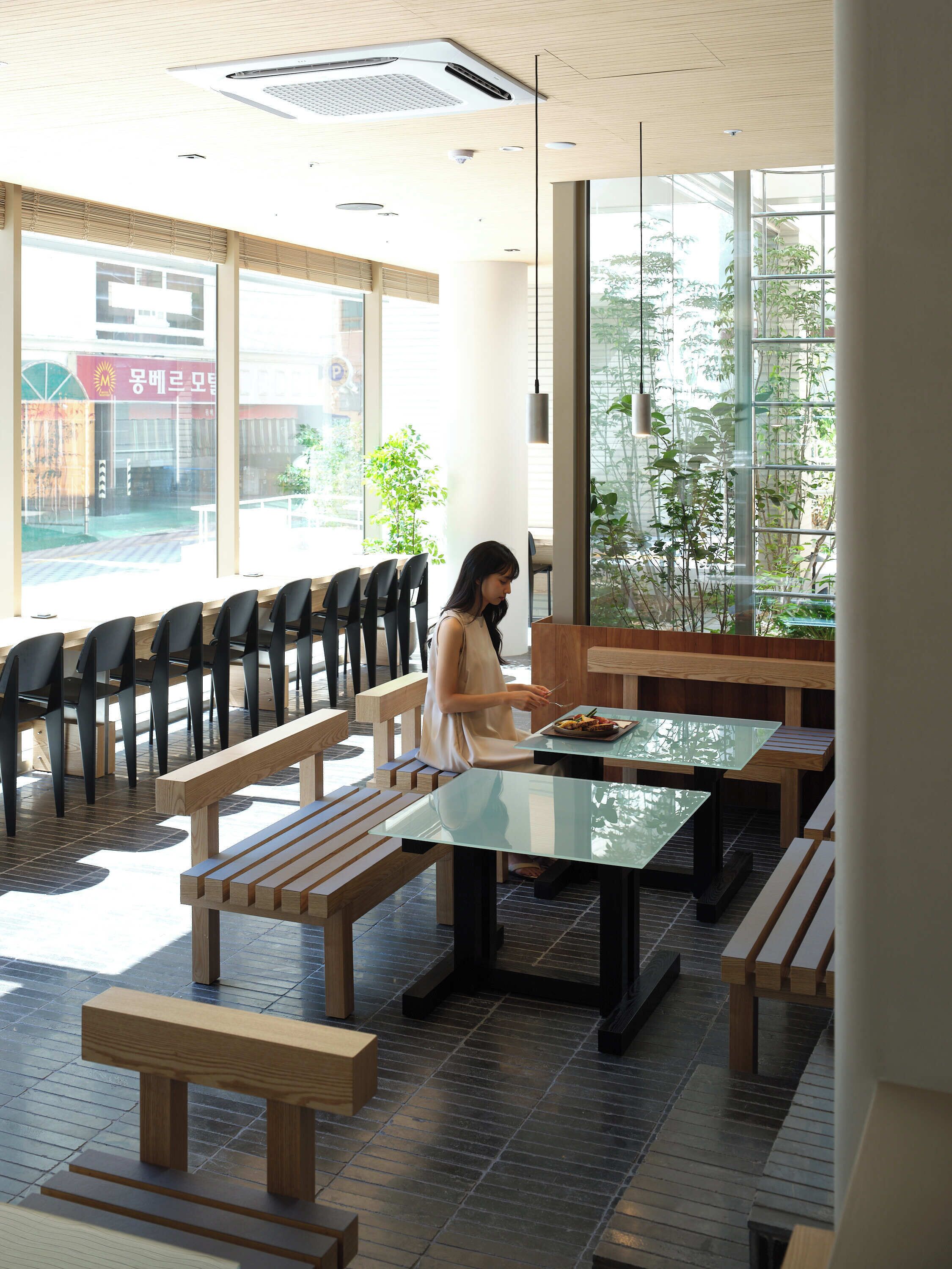Un interior moderno con bancos y mesas de madera, ventanas amplias y plantas de interior.