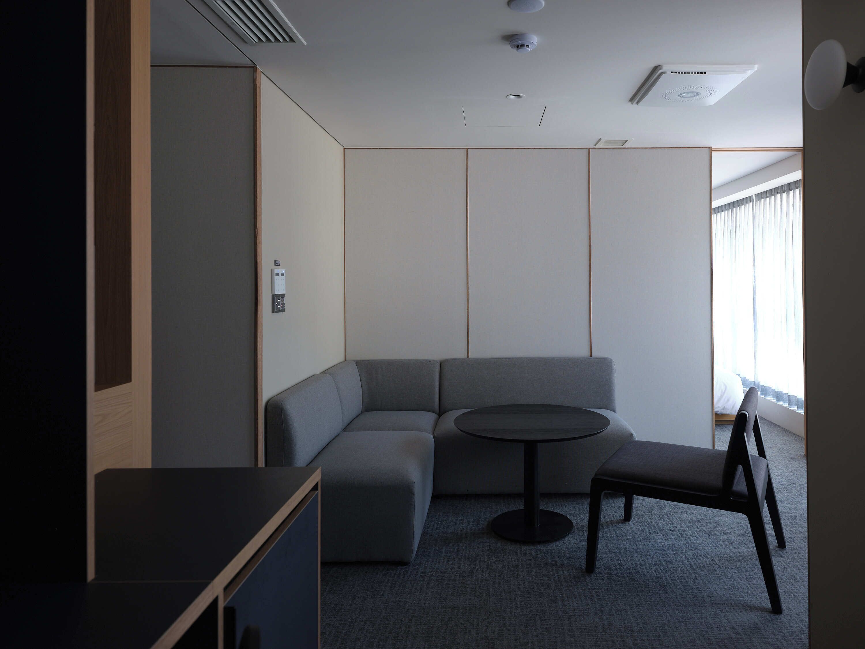 Una habitación moderna con un sofá modular, una mesita redonda y una silla. Hay una ventana con cortinas, un respiradero en el techo y una lámpara fijada a la pared.