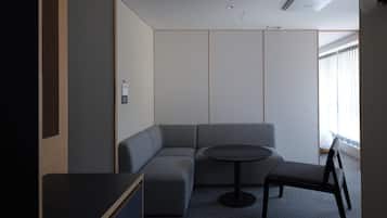 Una habitación moderna con un sofá modular, una mesita redonda y una silla. Hay una ventana con cortinas, un respiradero en el techo y una lámpara fijada a la pared.