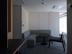 Una habitación moderna con un sofá modular, una mesita redonda y una silla. Hay una ventana con cortinas, un respiradero en el techo y una lámpara fijada a la pared.