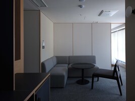 Una habitación moderna con un sofá modular, una mesita redonda y una silla. Hay una ventana con cortinas, un respiradero en el techo y una lámpara fijada a la pared.