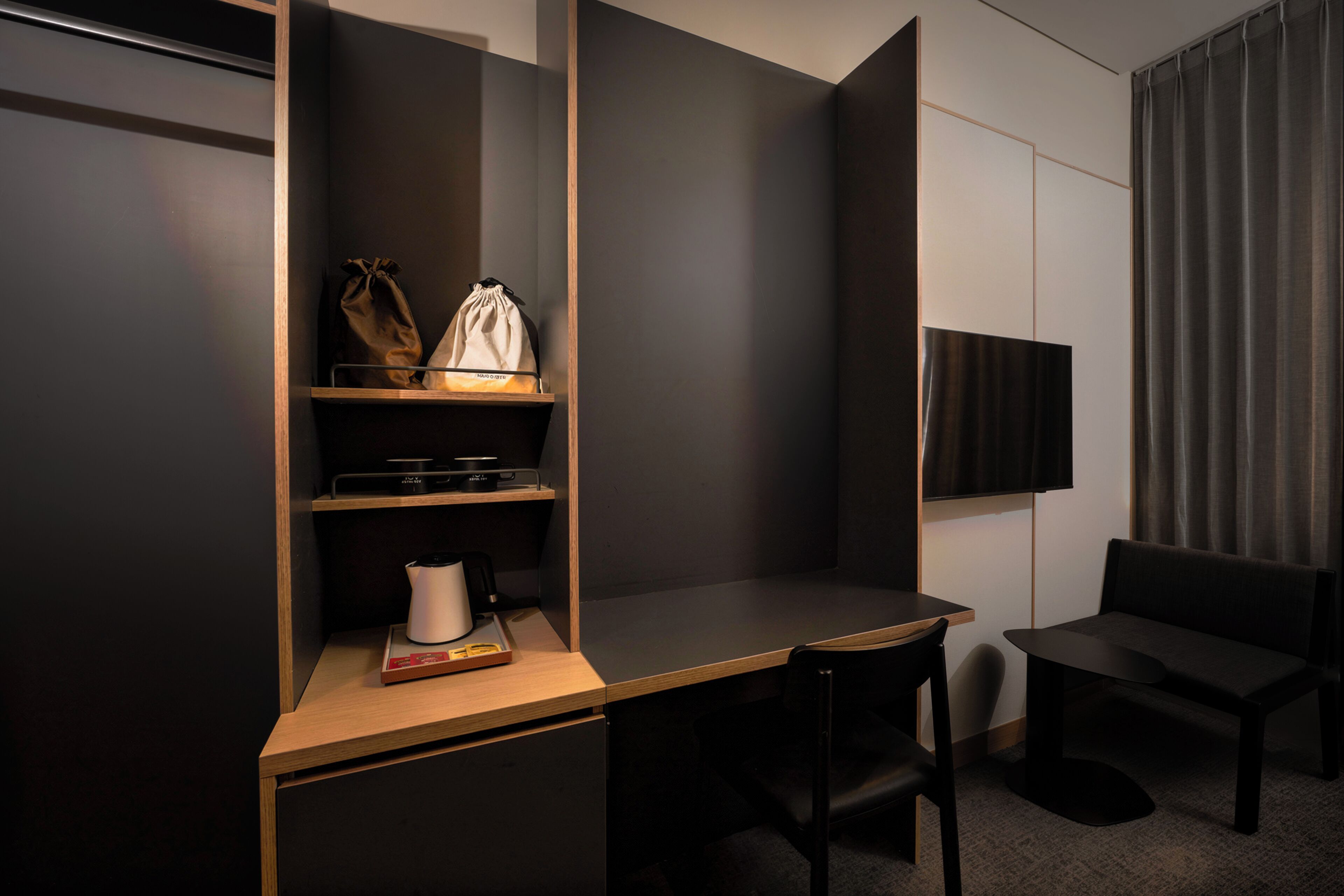 A modern hotel room with a desk, chairs, a TV, and a shelf with items.