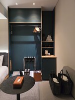Una habitación de hotel moderna con una cama, una mesa redonda con una caja de pañuelos, una zona de estar con un sofá, un armario con ropa colgada y un estante con una lámpara y un bolso.
