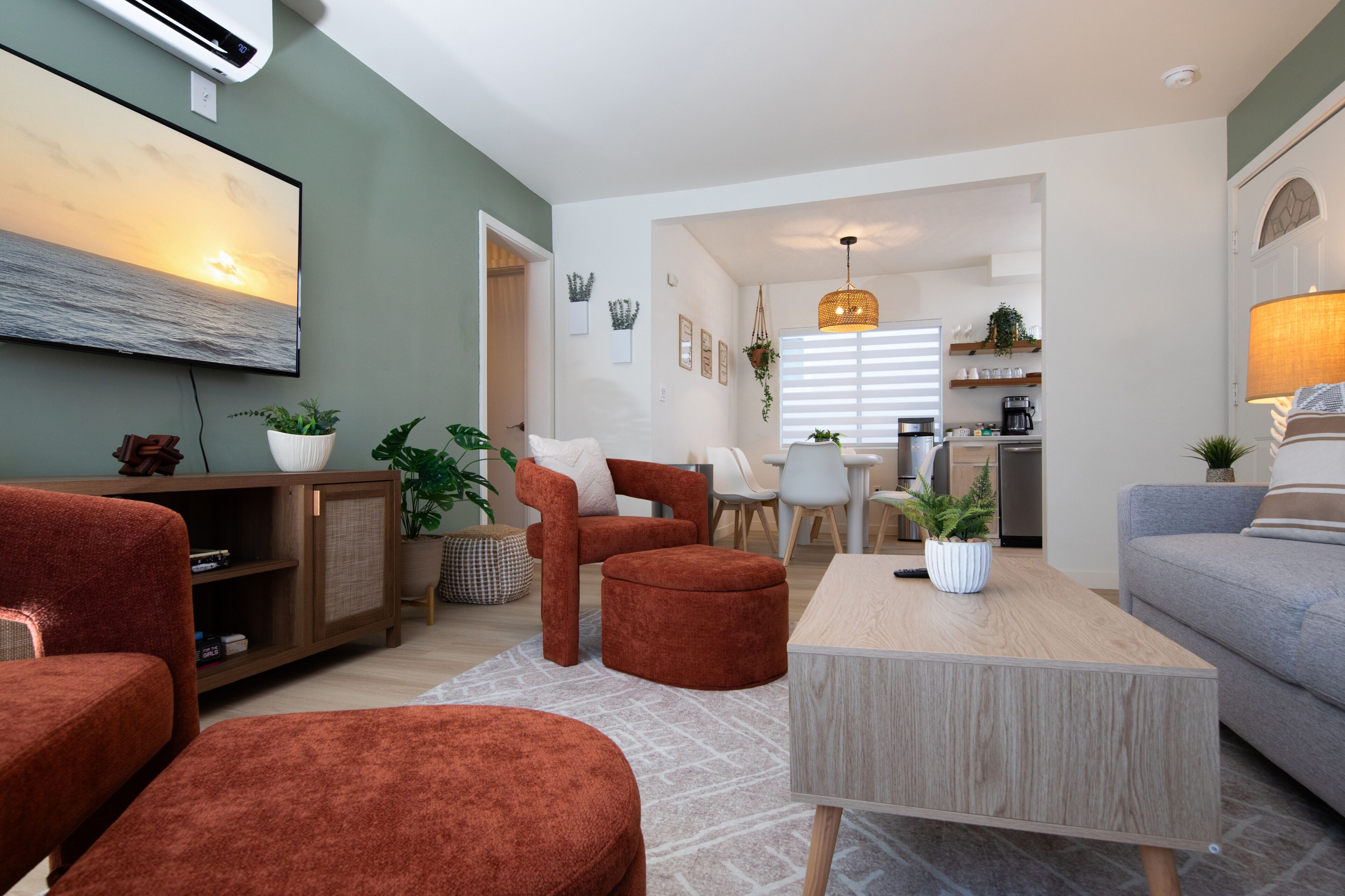 A modern living room with a sofa, ottoman, and coffee table, a dining area with chairs, a kitchen with appliances, and a large TV mounted on the wall.