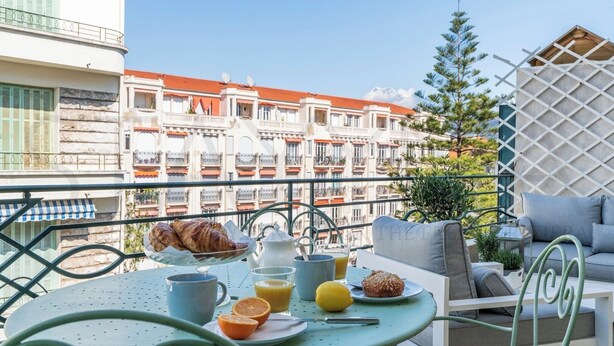 En balkon med et bord dækket til morgenmad, inklusive croissanter, appelsinjuice og en muffin, med udsigt over en række bygninger med balkoner.