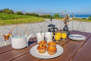 A breakfast setup with a teapot, cups, a plate of muffins, and a coffee maker on a wooden table with a scenic view.