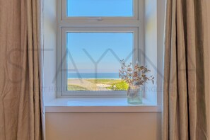 Ein Fenster mit Blick auf Strand und Meer, eine Vase mit getrockneten Blumen und Vorhänge.