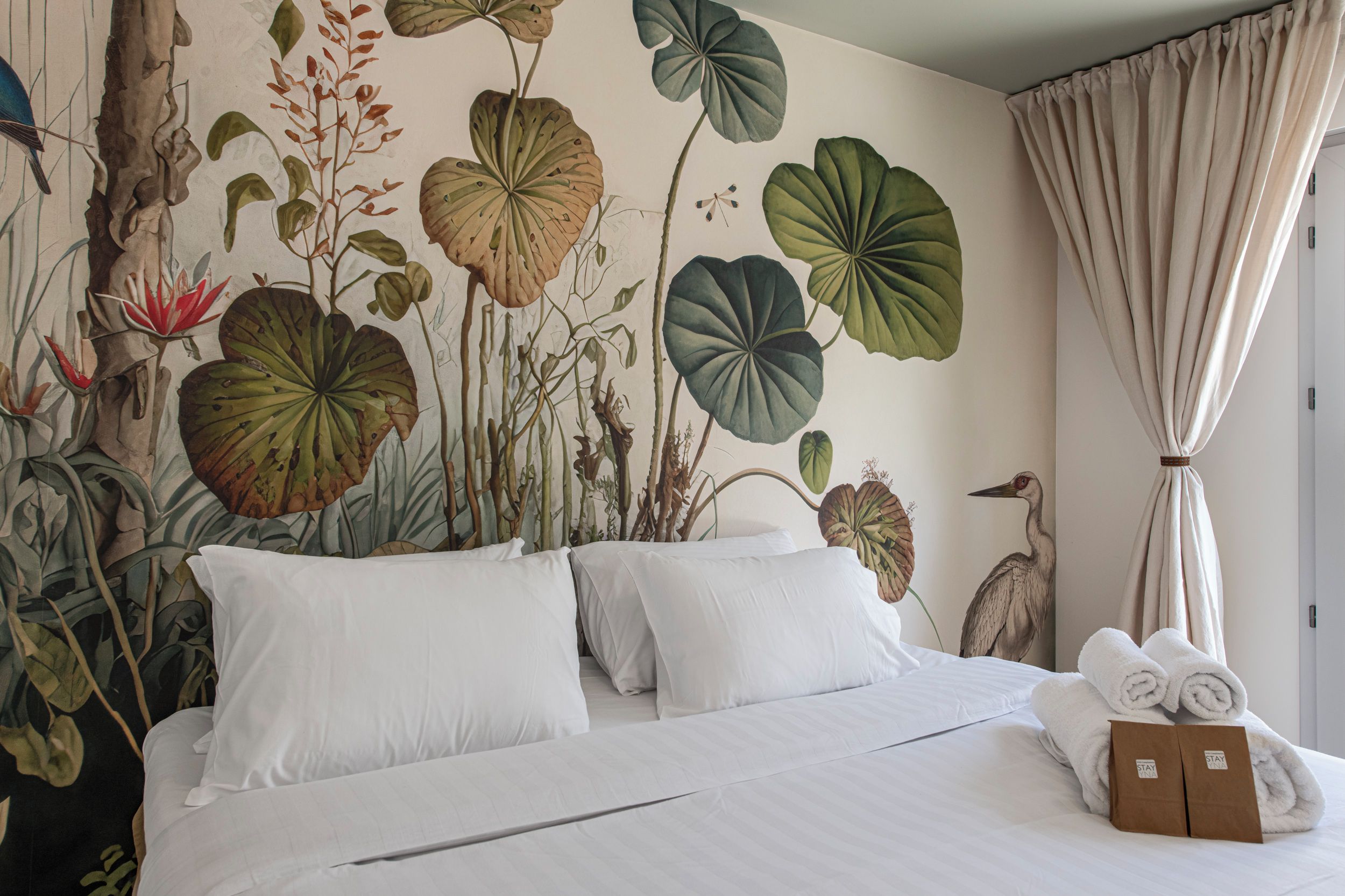 Ein Hotelzimmer mit einem großen Bett, weißer Bettwäsche, einem tropischen Wandbild und einem Vorhang.