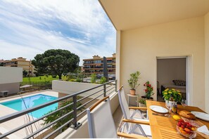 Un balcón con piscina, una mesa de comedor con frutas y una copa de vino, y vista a una zona residencial con árboles y edificios.