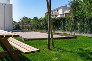 Un espacio al aire libre con césped, una banca de madera, un arenero pequeño y una cerca de metal.