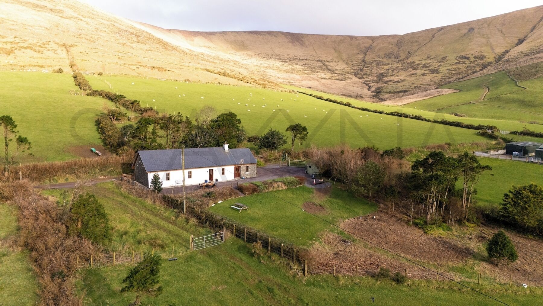 A rural house with a fenced yard, surrounded by green fields and hills.