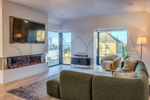 Ein modernes Wohnzimmer mit Kamin, Flachbildfernseher, einer Couch und Blick durch große Fenster.