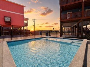 A swimming pool with a sunset in the background, surrounded by a fence and buildings. - CL5 Shared Pool, Charcoal Grill, Old Town Port A (Port Aransas)