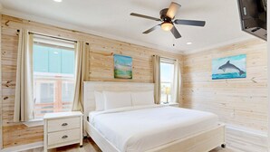 A bedroom with a bed, nightstand, ceiling fan, wooden walls, and artwork. - CL5 Shared Pool, Charcoal Grill, Old Town Port A (Port Aransas)