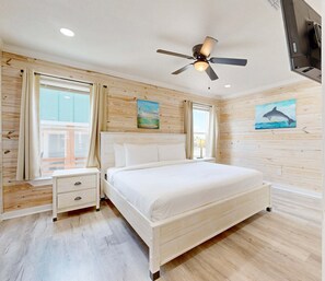 A bedroom with a bed, nightstand, ceiling fan, wooden walls, and artwork. - CL5 Shared Pool, Charcoal Grill, Old Town Port A (Port Aransas)