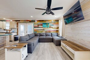 A modern living room with a sectional sofa, a ceiling fan, a flat-screen TV, and a colorful wooden wall. - CL5 Shared Pool, Charcoal Grill, Old Town Port A (Port Aransas)