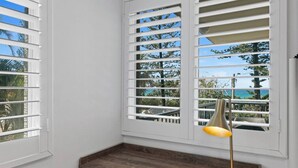 Una habitación de esquina con paredes blancas, un escritorio de madera y una ventana con contraventanas blancas.
