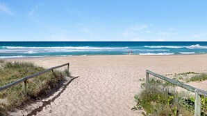 Una playa con un camino de madera que lleva al agua, arena dorada y dos personas caminando.