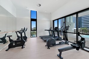 A modern gym with cardio machines, a large window, and a view of buildings. - Oceanview Getaway | 2B Apartment. Pool. Parking (Mermaid Beach)
