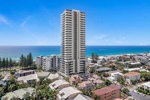 A tall, modern hotel with a central atrium, surrounded by residential buildings and a coastal view. - Oceanview Getaway | 2B Apartment. Pool. Parking (Mermaid Beach)