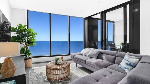 Ein modernes Wohnzimmer mit einem grauen Sofa, einem runden Kaffeetisch aus Rattan und Blick auf das Meer.